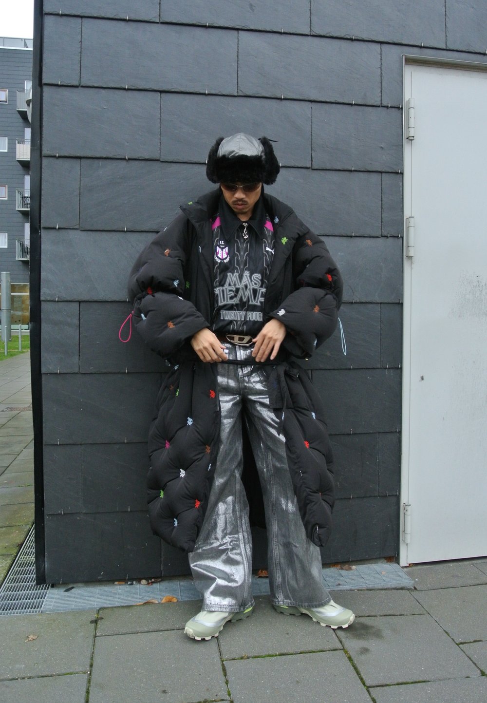 Cappotto nero imbottito con ricami multicolore, pantaloni argentati a gamba larga e sneakers verdi chunky, abbinati a un cappello di pelliccia e una t-shirt grafica.