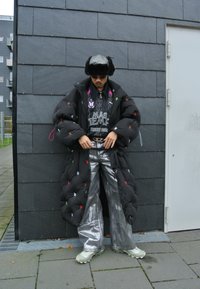 Cappotto nero imbottito con ricami multicolore, pantaloni argentati a gamba larga e sneakers verdi chunky, abbinati a un cappello di pelliccia e una t-shirt grafica.