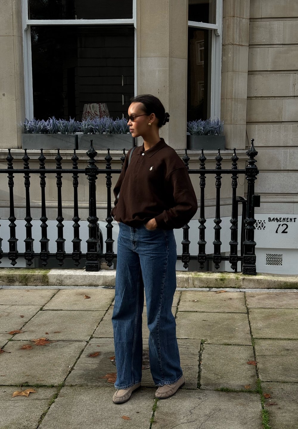 Polo à manches longues marron avec un petit logo, jean denim bleu à jambes larges, et chaussures grises sans lacets. Debout contre une rambarde noire.