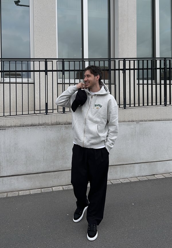 Lächelnder Mann in grauem Kapuzenpullover mit Reißverschluss, schwarzer Hose und Turnschuhen, der eine schwarze Mütze hält und auf dem Gehsteig neben einem Steingebäude mit Fenstern und Geländer steht.