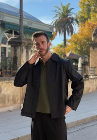 Homme en veste noire et chemise vert olive debout dans une rue ensoleillée avec un palmier et un vieux bâtiment en pierre en arrière-plan.