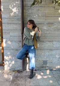 Sweatshirt verde, jeans azul claro e botas de tornozelo pretas. O fundo apresenta pedra texturizada e um tubo marrom vertical.