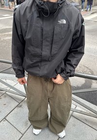 Άτομο που φοράει μαύρο μπουφάν North Face, χαλαρά παντελόνια cargo σε χακί απόχρωση και λευκά αθλητικά παπούτσια, στέκεται σε πεζοδρόμιο πόλης κοντά σε μεταλλική περίφραξη.