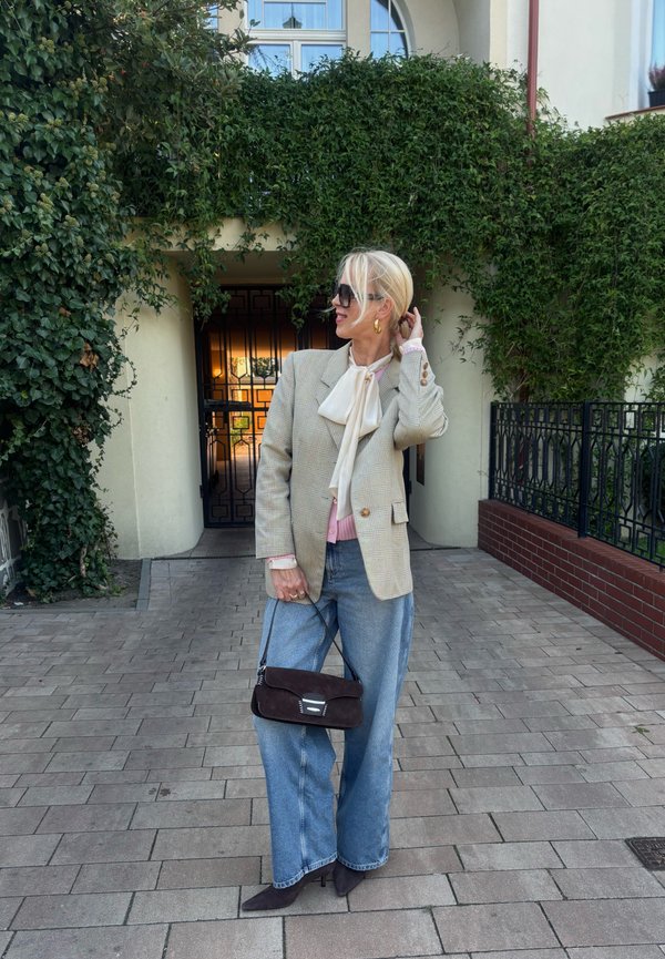 Blazer a quadri beige, blusa rosa chiaro con fiocco, jeans larghi blu, borsa in suede marrone scuro e occhiali da sole neri. Viale pavimentato con vegetazione.