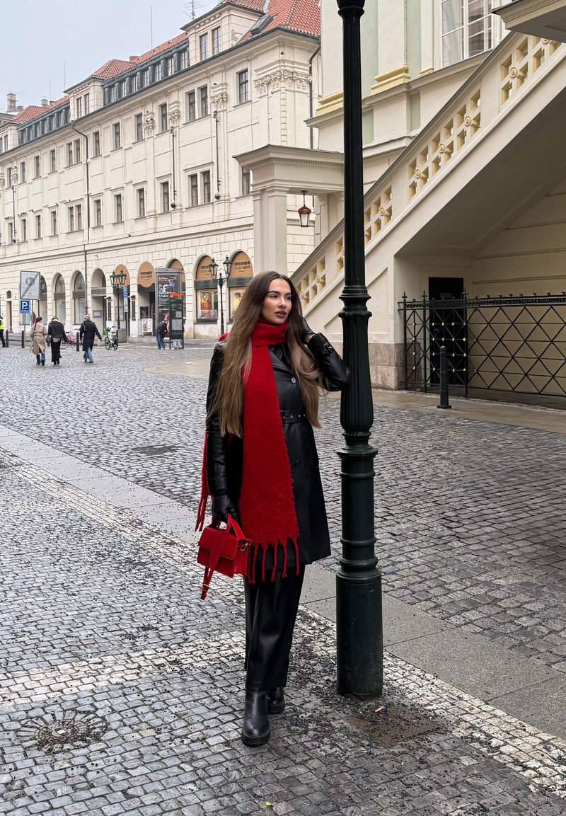 Woman in black leather coat and boots with red scarf and bag standing by a lamppost on a cobblestone street near historic buildings.