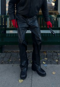 Persona vestida con pantalones de cuero negro, botas negras y guantes rojos, sosteniendo gafas de sol negras, de pie en una acera cerca de un banco verde.