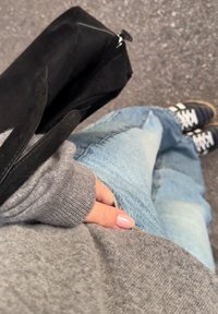 Personne portant un pull gris et un jean bleu délavé, tenant un sac à main noir, debout sur un trottoir gris avec des baskets noires.