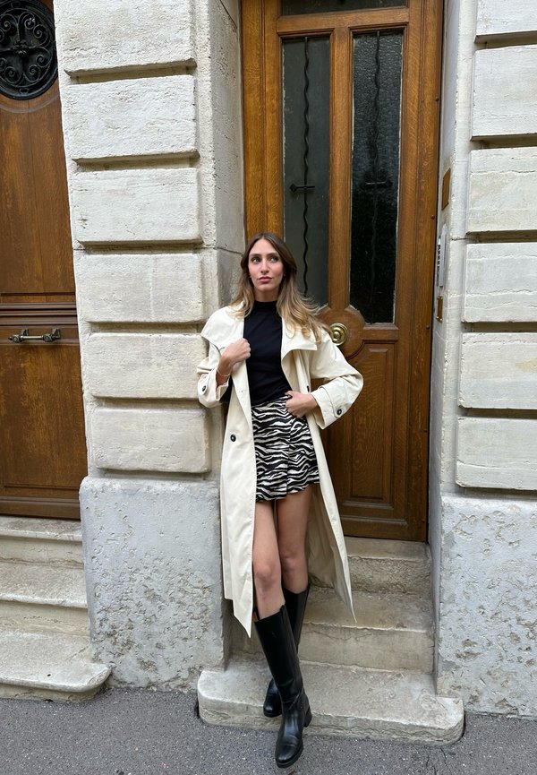 Mujer con un abrigo largo beige, top negro, falda con estampado de cebra y botas negras apoyada contra una pared de piedra cerca de una puerta de madera.