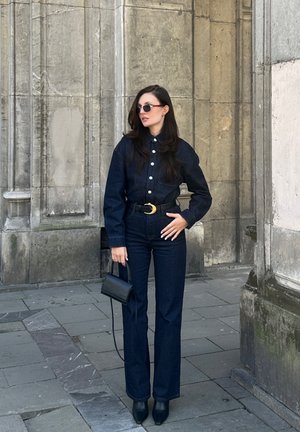 Conjunto de denim oscuro que incluye una camisa de botones con un bolsillo y pantalones acampanados, combinados con tacones negros de punta y gafas de sol. Pequeño bolso negro.