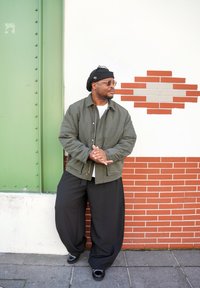 Homme en veste verte, pantalon large noir, béret noir et lunettes de soleil, se tient contre un mur blanc en briques avec un panneau métallique vert.