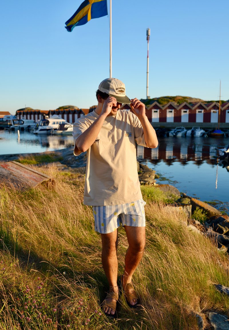 Man adjusts cap standing on grassy shore near boats and red boathouses with Swedish flag and clear sky in background.