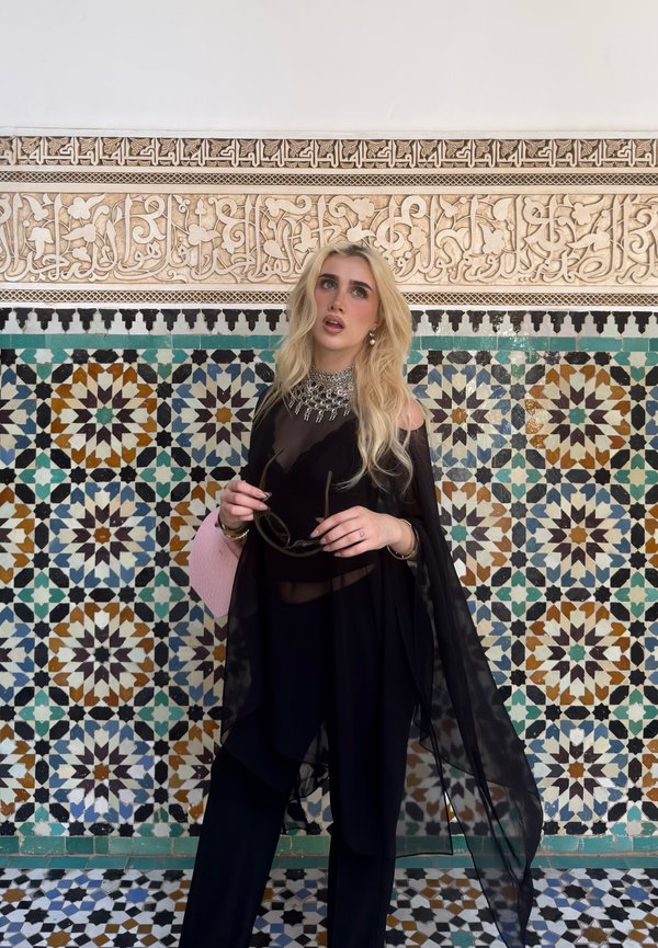 Donna bionda con abito nero trasparente e collana vistosa in piedi che tiene occhiali da sole di fronte a una parete colorata con piastrelle geometriche e un pannello intagliato ornato.