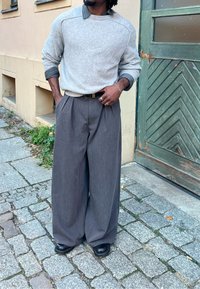 Svetlosivý pletený sveter s rebrovaným golierom, vrstvený cez tmavú košeľu s golierom; široké sivé plisované nohavice, čierne topánky a čierny opasok.