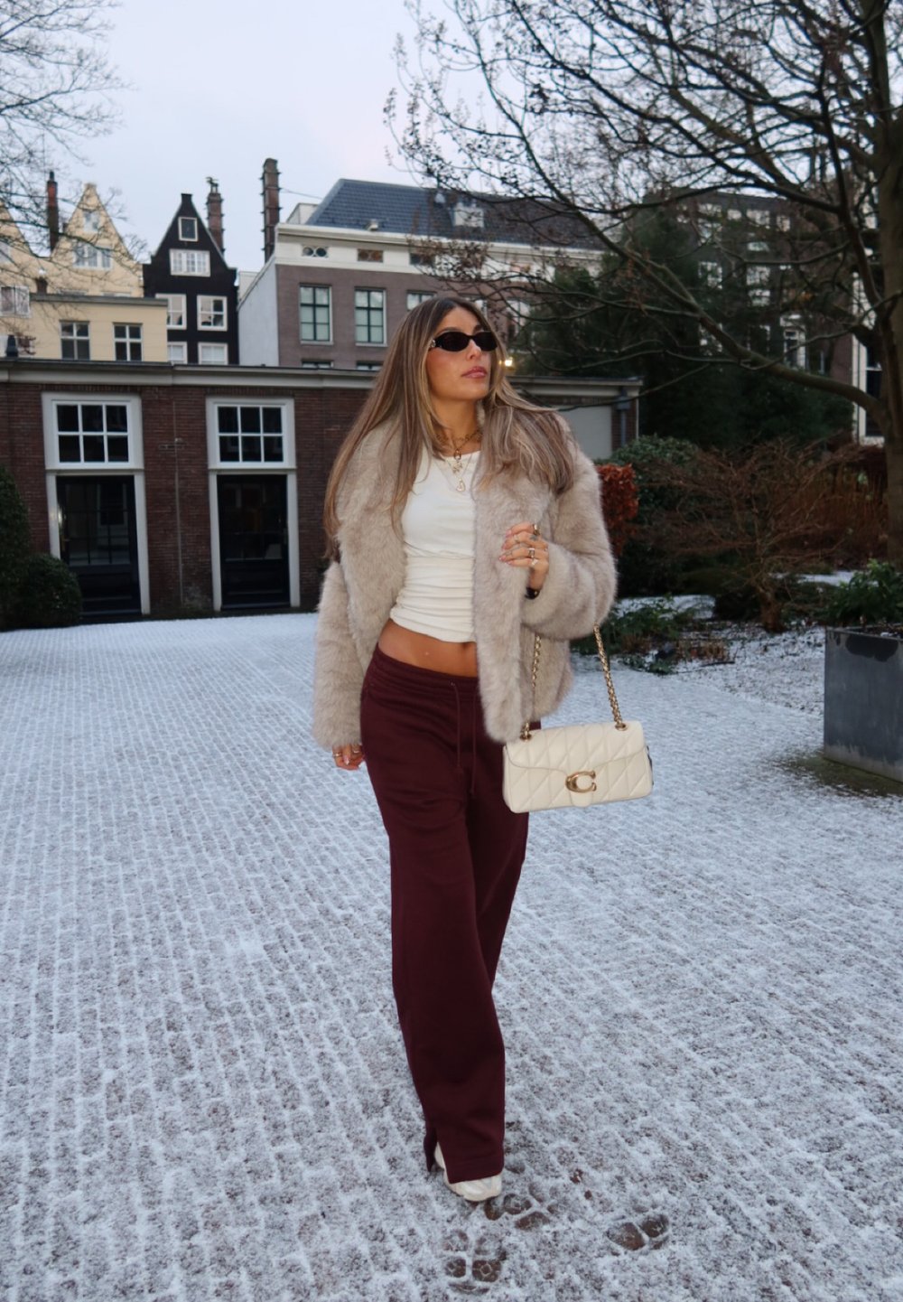 Γυναίκα που φοράει γούνινο πανωφόρι, λευκό cropped τοπ και μπορντό παντελόνι περπατάει πάνω σε χιονισμένα λιθόστρωτα δρόμο κρατώντας μια κρεμ τσάντα σε αστικό περιβάλλον.