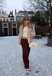 Γυναίκα που φοράει γούνινο πανωφόρι, λευκό cropped τοπ και μπορντό παντελόνι περπατάει πάνω σε χιονισμένα λιθόστρωτα δρόμο κρατώντας μια κρεμ τσάντα σε αστικό περιβάλλον.