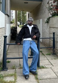 Casaco insulated preto encurtado sobre uma t-shirt branca, combinado com jeans de denim azul largos e sapatos pretos. Gorro cinza com padrão e óculos.