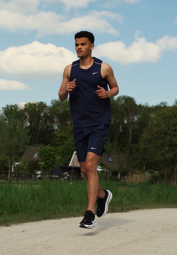 Joven vestido con ropa deportiva azul corriendo por un camino de tierra con árboles y casas al fondo bajo un cielo parcialmente nublado.
