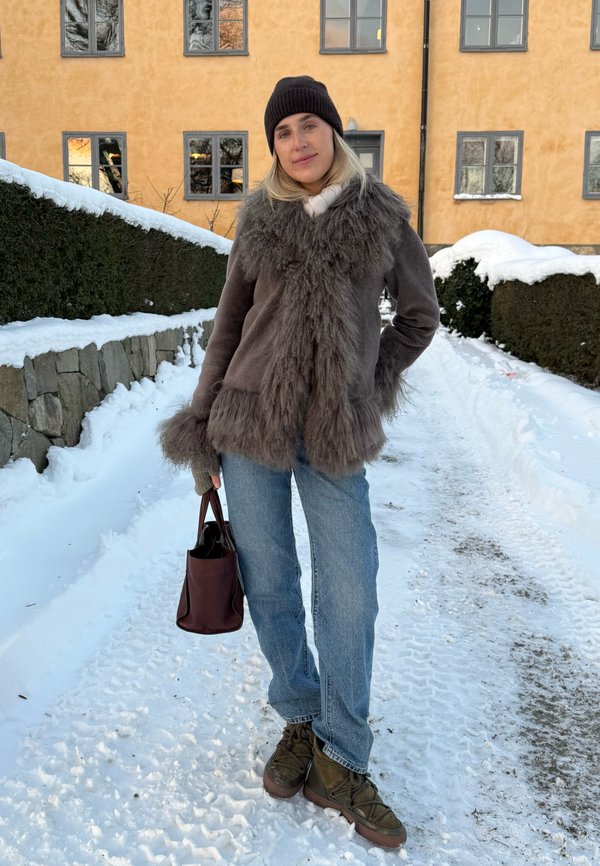 Nő meleg szőrmekabátban, sapkában, farmerben és csizmában, barna táskát tartva, egy havas ösvényen áll, mögötte sárga épület és sövények.