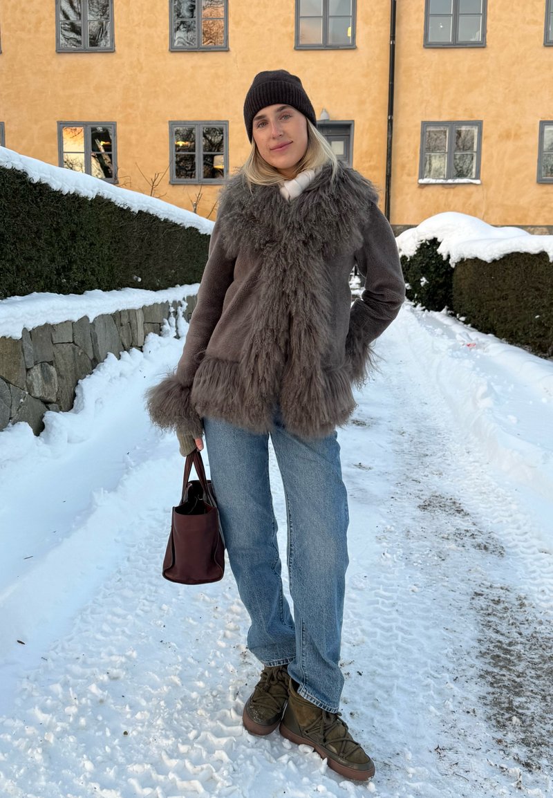 Femme en manteau de fourrure chaud, bonnet, jean et bottes, tenant un sac marron, debout sur un chemin enneigé avec un bâtiment jaune et des haies en arrière-plan.