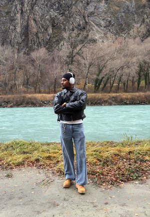 Chaqueta bomber de cuero negro, jeans anchos azul claro y zuecos naranjas; lleva auriculares blancos y un gorro negro cerca de la orilla del río.