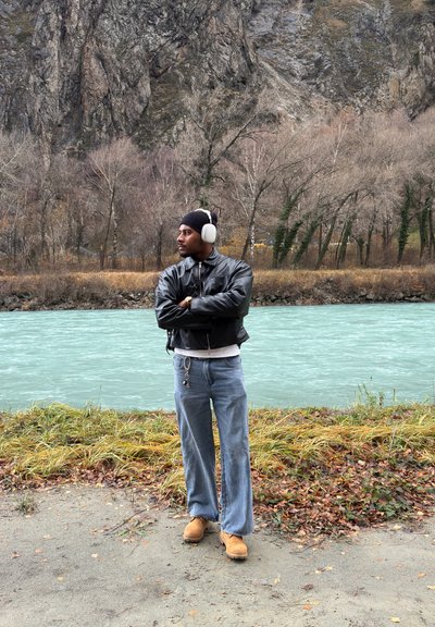 Chaqueta bomber de cuero negro, jeans anchos azul claro y zuecos naranjas; lleva auriculares blancos y un gorro negro cerca de la orilla del río.