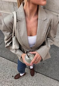 Lichtgrijze geruite blazer met een riem, witte top, blauwe jeans en brown schoenen. Houdt een transparante beker met een rietje vast voor een gestructureerde achtergrond.