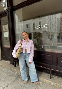 Gestreepte, korte, lange mouwen blouse in lichtroze; losse lichtblauwe jeans; beige schoenen; geweven tas met een bloemdetail; weerspiegeling in het raam.