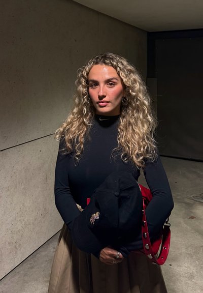 Gorra negra con bordado sostenida en los brazos, combinada con una blusa negra de manga larga ajustada y una falda plisada beige. Destaca un bolso rojo como acento.