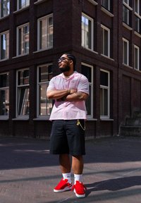 Camisa cor-de-rosa de manga curta com gola, calções pretos e ténis vermelhos. Apresenta óculos de sol e uma corrente decorativa presa aos calções. Fundo em tijolo.