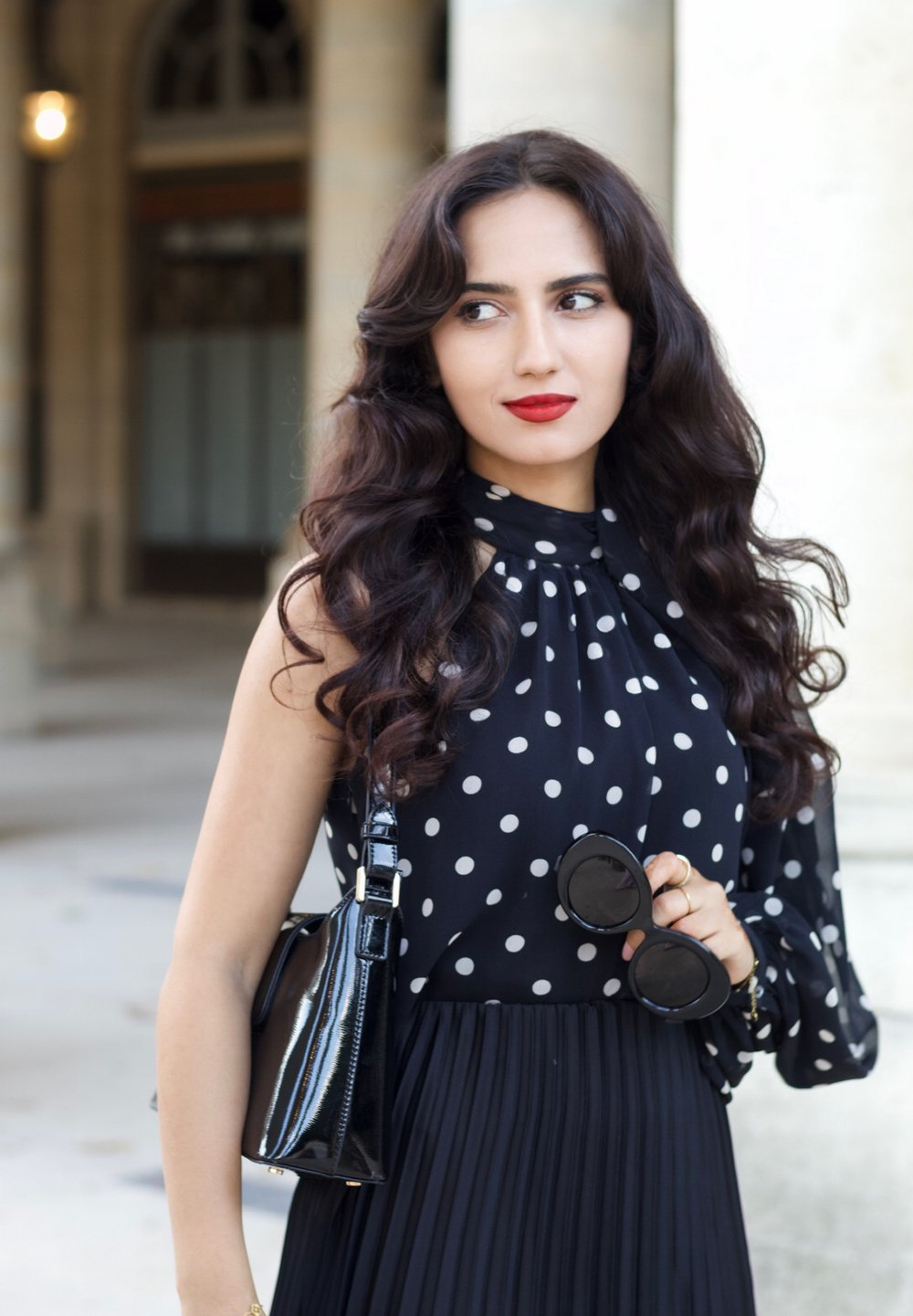 Blusa nera a pois con collo alto, abbinata a una gonna nera plissettata. Porta occhiali da sole neri rotondi e tiene una borsa nera piccola.