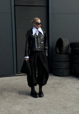 Persona con chaqueta de cuero negra con cuello blanco grande, pantalones anchos negros, botas de combate, gafas de sol y sosteniendo una bolsa negra cerca de neumáticos apilados.