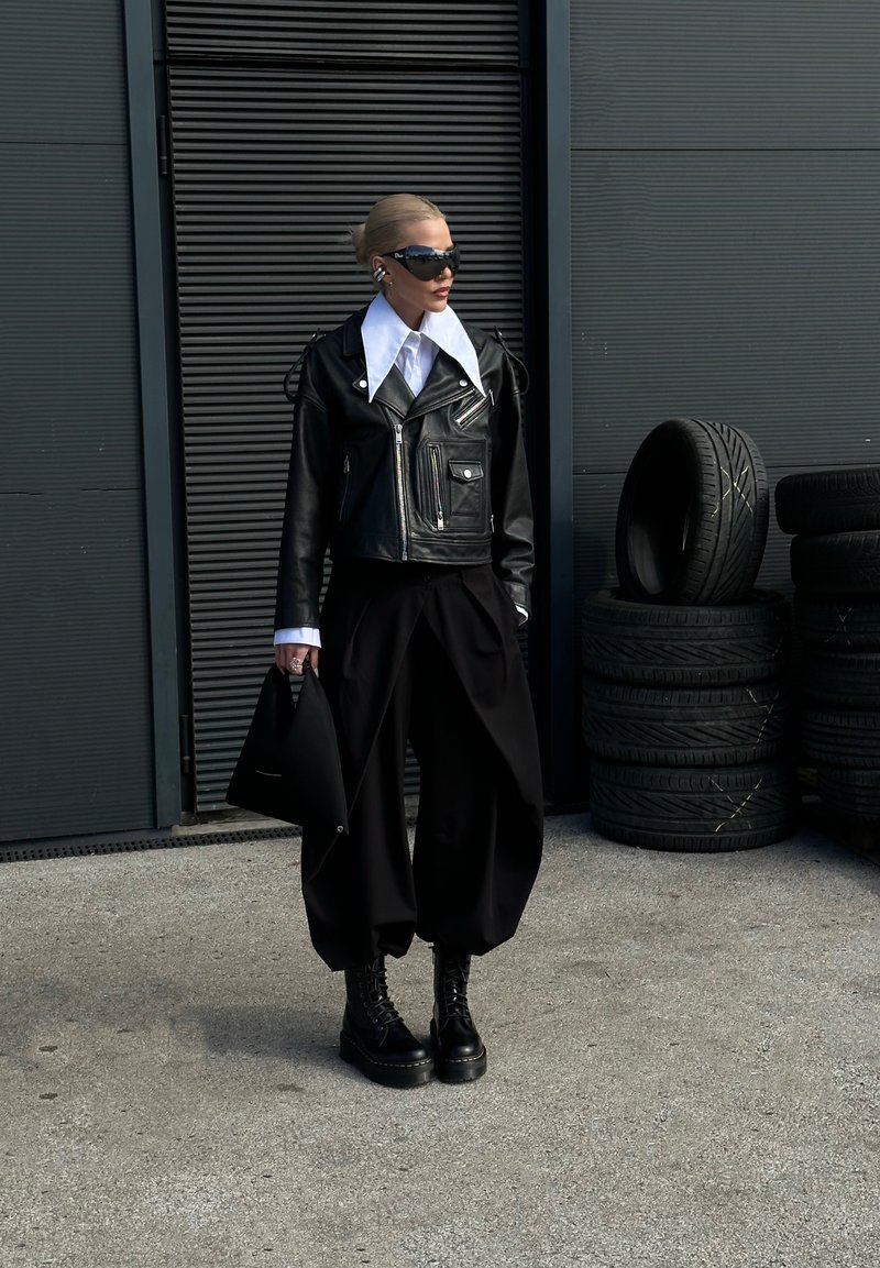 Personne portant une veste en cuir noire avec un col blanc oversize, un pantalon large noir, des bottes de combat, des lunettes de soleil, et tenant un sac noir près de pneus empilés.