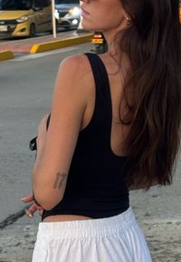 Zwarte rugloze tanktop, witte shorts, arm met een tattoo. Lang, donker haar valt over de schouder. Straat en voertuigen op de achtergrond.