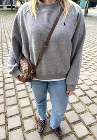 Personne portant un sweat-shirt gris, un jean bleu clair, des chaussures en cuir marron et un sac bandoulière avec un motif beige et marron, debout sur un chemin pavé.