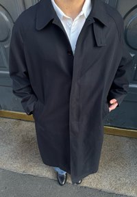 Personne portant un long manteau noir sur une chemise blanche et un jean bleu, les mains dans les poches du manteau, debout sur une surface en pierre et en asphalte.