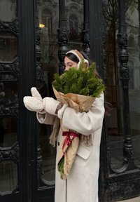 Femme en manteau beige et moufles tenant un bouquet de branches de pin enveloppé de papier avec un ruban rouge, debout près d'une porte noire ornée.