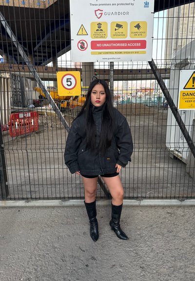 Chaqueta de tela negra de gran tamaño con un alto cuello, combinada con pantalones cortos negros y botas de rodilla de punta. En un fondo de construcción.