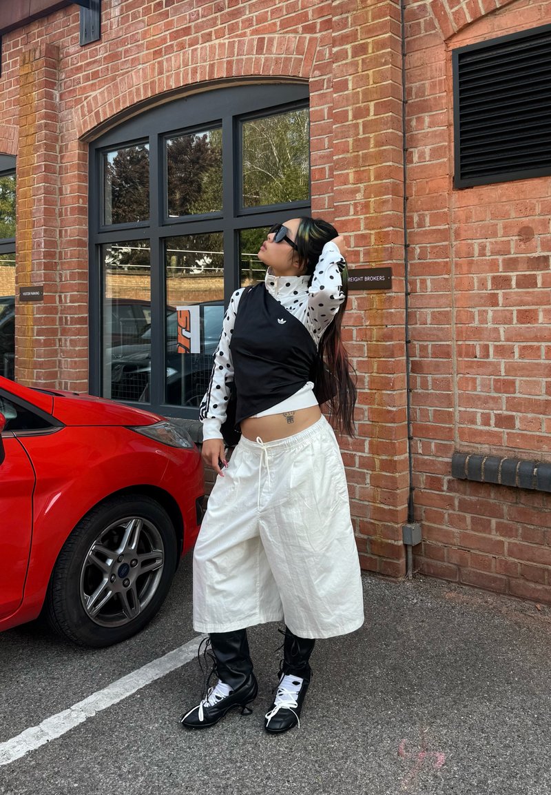 Personne portant une tenue noire et blanche avec des lunettes de soleil se tient près d'une voiture rouge devant un bâtiment en briques avec de grandes fenêtres.