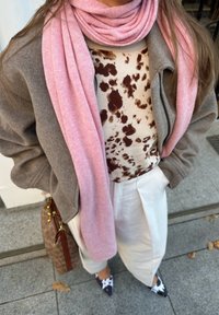 Giacca grigia oversize, top beige con macchie marroni, sciarpa rosa pallido, pantaloni bianchi a pieghe e scarpe scure con motivo. Borsa marrone visibile.