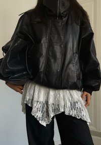 Svart skinnjakke i stor størrelse med høy krage og glidelås, lagd over en hvit blondesbluse med bølget kant, brukt sammen med svarte bukser.