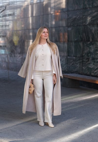 Mujer con cabello largo y rubio que lleva una blusa crema, pantalones y abrigo, sosteniendo un bolso pequeño beige, de pie sobre una acera urbana cerca de una pared de piedra oscura.