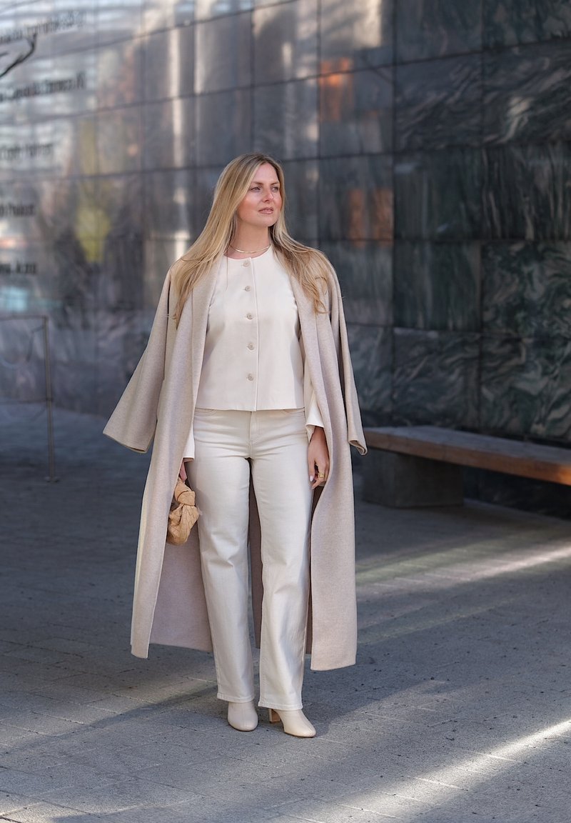 Femme aux longs cheveux blonds portant un chemisier, un pantalon et un manteau crème, tenant un petit sac beige, debout sur un trottoir urbain près d'un mur en pierre sombre.