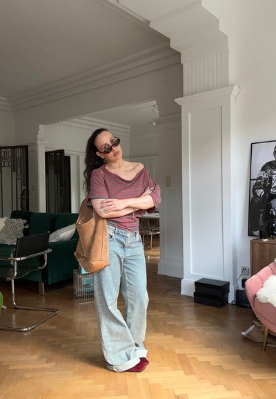 Mujer con gafas de sol, top rojo a rayas sin hombros, vaqueros anchos y zapatos rojos, de pie en un salón elegante con suelo de madera y muebles modernos.
