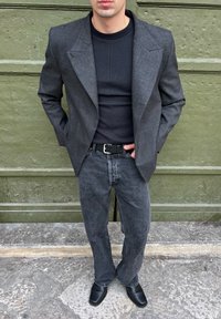 Blazer gris texturizado sobre una camiseta ajustada negra, combinada con jeans de color gris oscuro y zapatos de cuero negros. El conjunto incluye un cinturón negro.