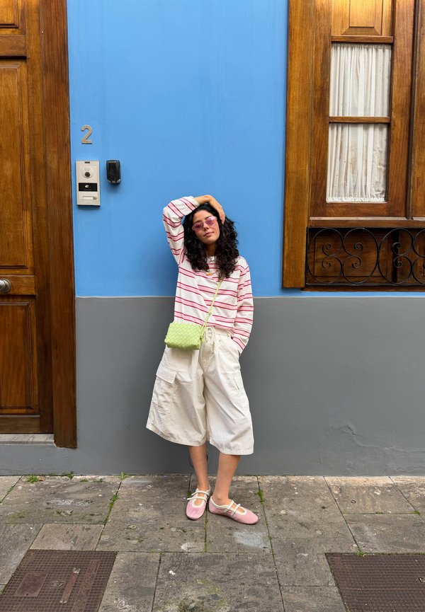 Femme aux cheveux bouclés, portant une chemise à rayures roses, un short, des chaussures roses et un sac vert, appuyée contre un mur bleu-gris près d'une porte en bois et d'une fenêtre.