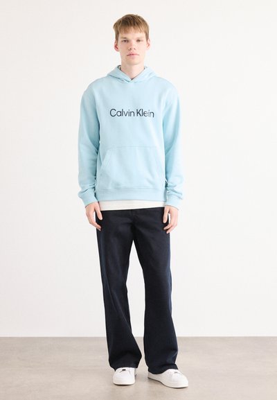 Sudadera con capucha de color azul claro, con un bolsillo frontal y el logo de "Calvin Klein". Combinada con pantalones de navy oscuro y calzado blanco sobre un fondo neutro.