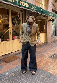 Άνδρας που φοράει aviator καπέλο με γούνα, ριγέ πουκάμισο με μοτίβο, καρό σακάκι, φαρδιά μαύρα παντελόνια και παπούτσ�ια με σχέδιο, στέκεται σε πεζοδρόμιο έξω από καφέ.