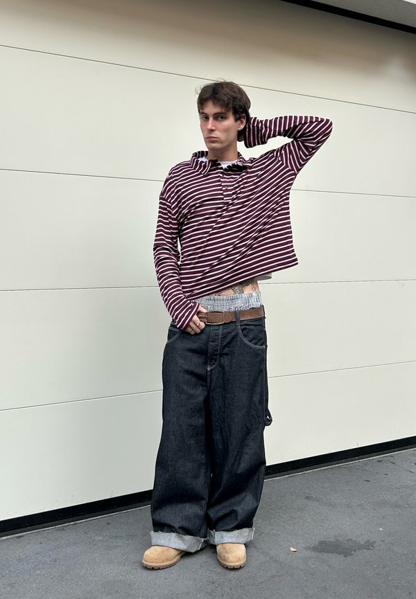 Maroon and white striped long-sleeve shirt, loose-fitting dark denim jeans with rolled cuffs, beige footwear, standing against a plain background.