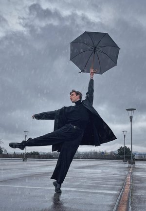 Individuo con un largo abrigo negro y un cuello de tortuga negro, sostiene un paraguas negro, de pie sobre un pavimento húmedo con una pose dramática contra un cielo gris.