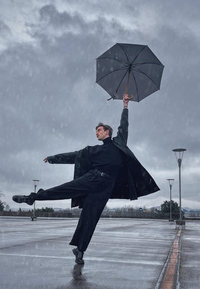 Individuo con un largo abrigo negro y un cuello de tortuga negro, sostiene un paraguas negro, de pie sobre un pavimento húmedo con una pose dramática contra un cielo gris.